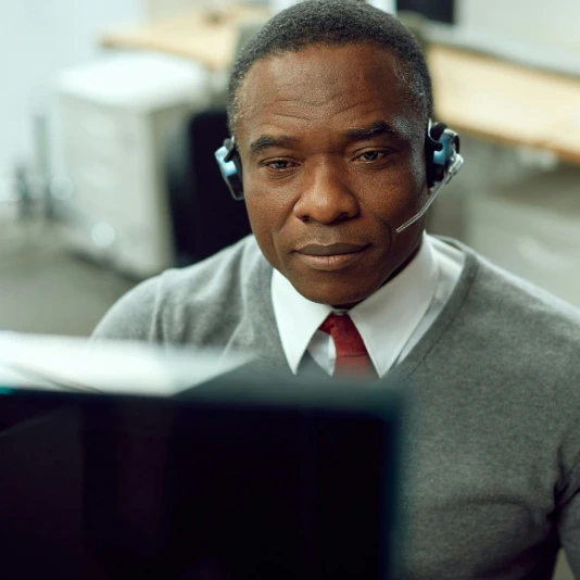 Man wearing a headset, gray sweater, and red tie, looking at a computer screen.