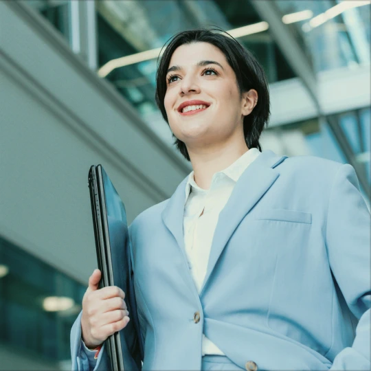A smiling woman in a blue suit, holding a laptop, stands in a modern office building.