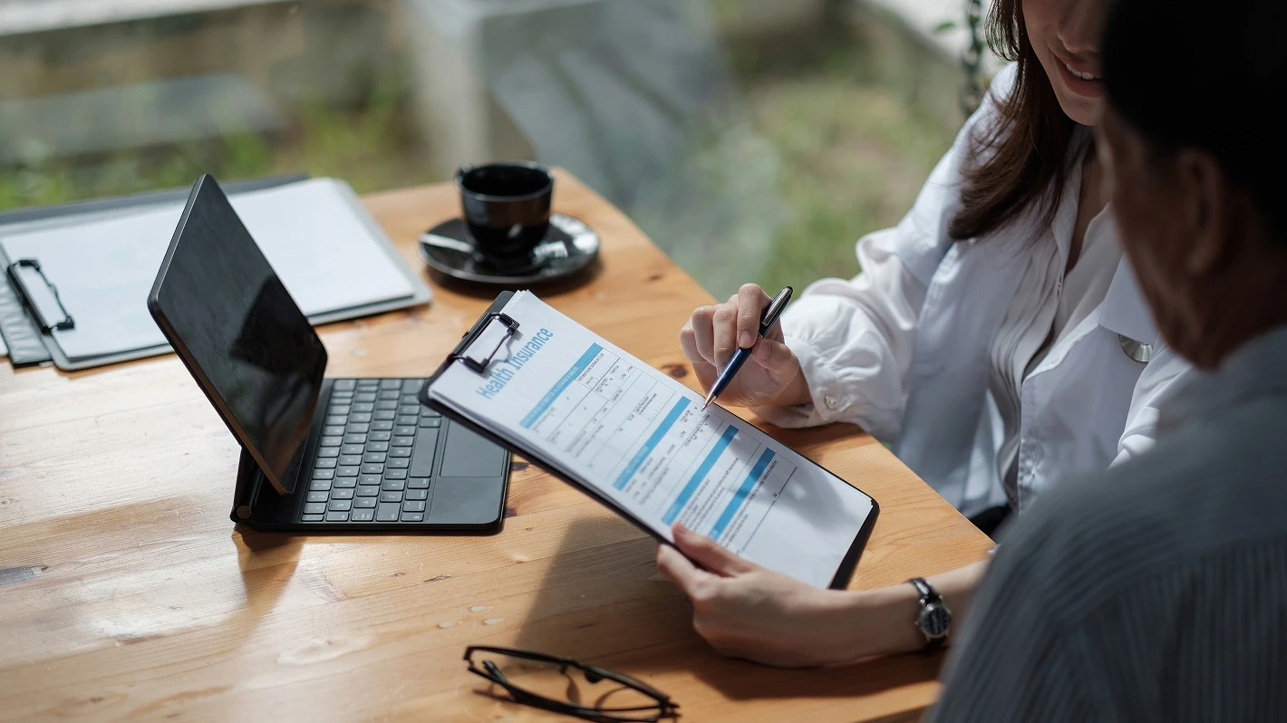 Close-up of a health insurance agent reviewing a form with a client at a desk with a laptop and coffee