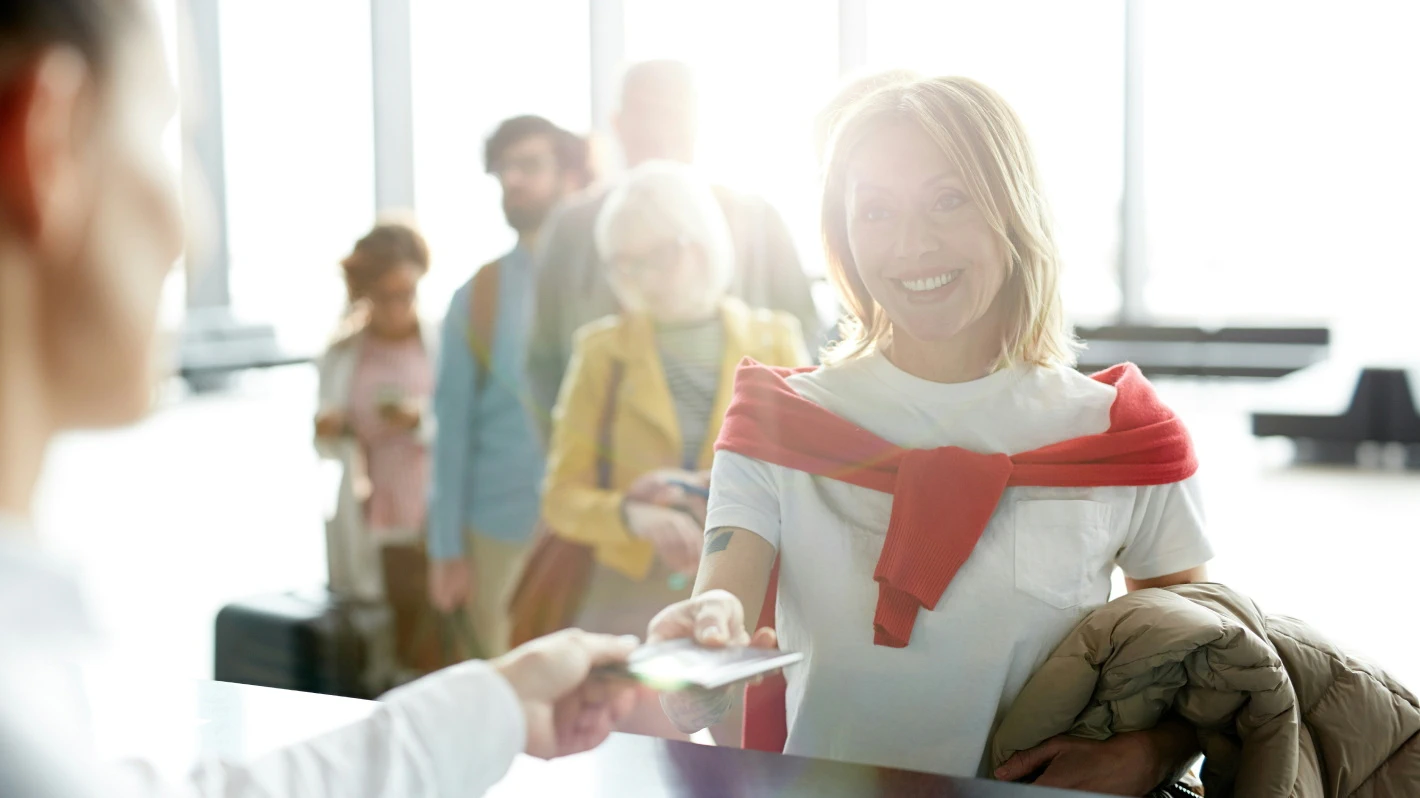 A smiling woman hands her ticket to an airport attendant, with a line of waiting passengers visible in the background