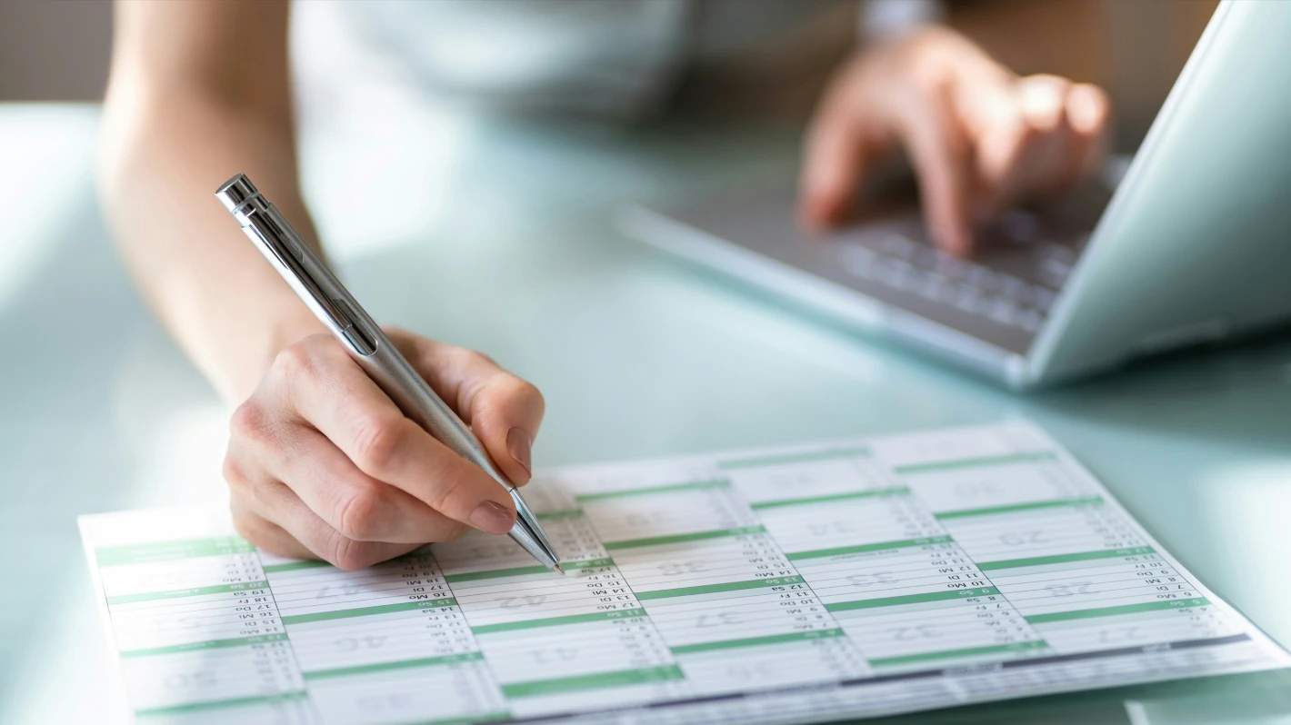 Close-up of a person's hands marking dates on a calendar while using a laptop for scheduling