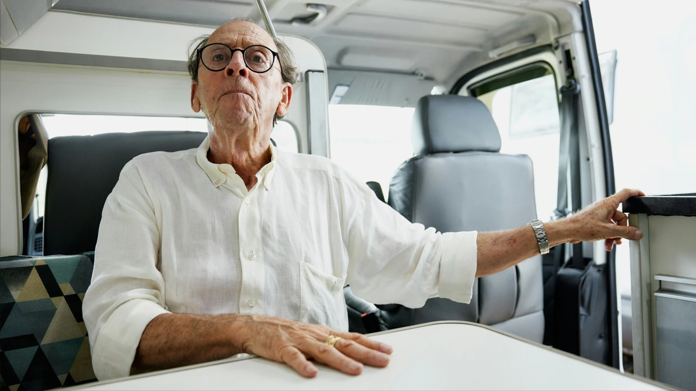 Older man with glasses, wearing a white linen shirt, sitting inside a van with his hand resting on a small table