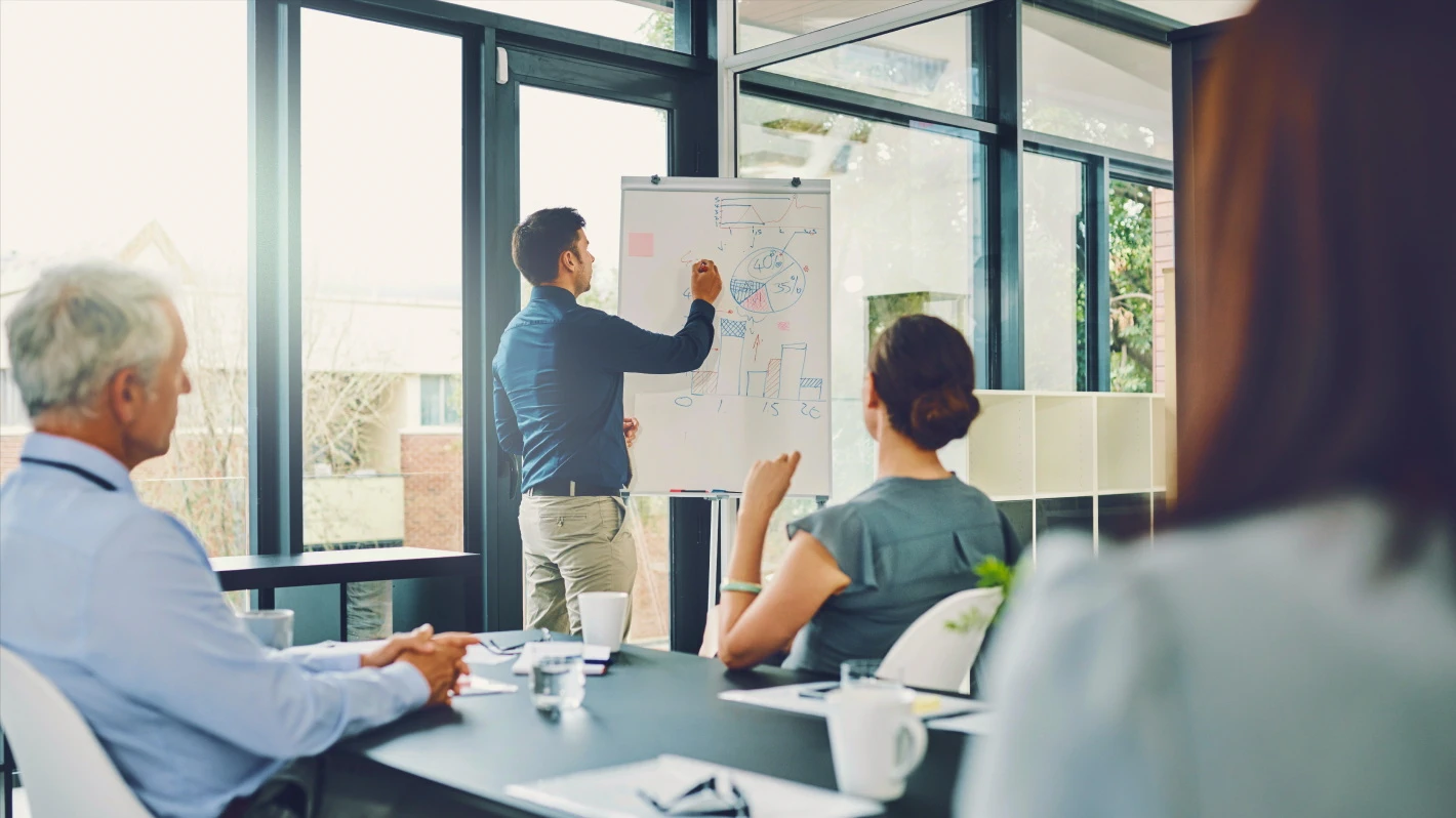 A man in a blue shirt is presenting to a group, drawing on a whiteboard that displays diagrams and data. The meeting takes place in a room with large windows.