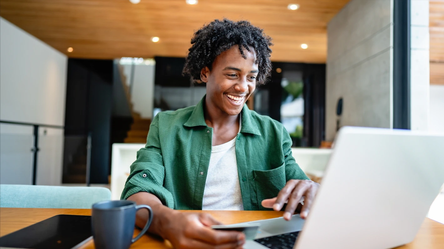 A smiling man wearing a green shirt sits at a table and uses a laptop, holding a credit card in his hand