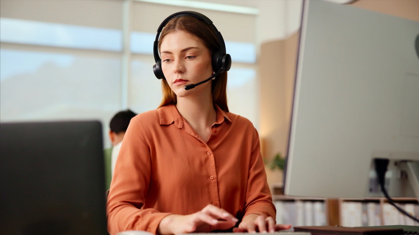 Focused female customer support agent with long red hair, wearing an orange blouse and a headset, working at a computer