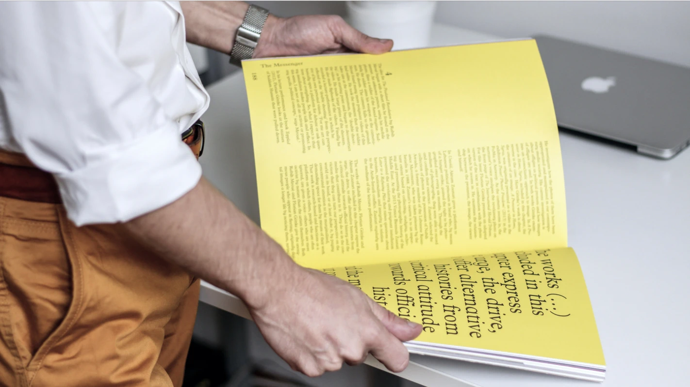 A man wearing orange pants and a white shirt holds open a book with yellow pages
