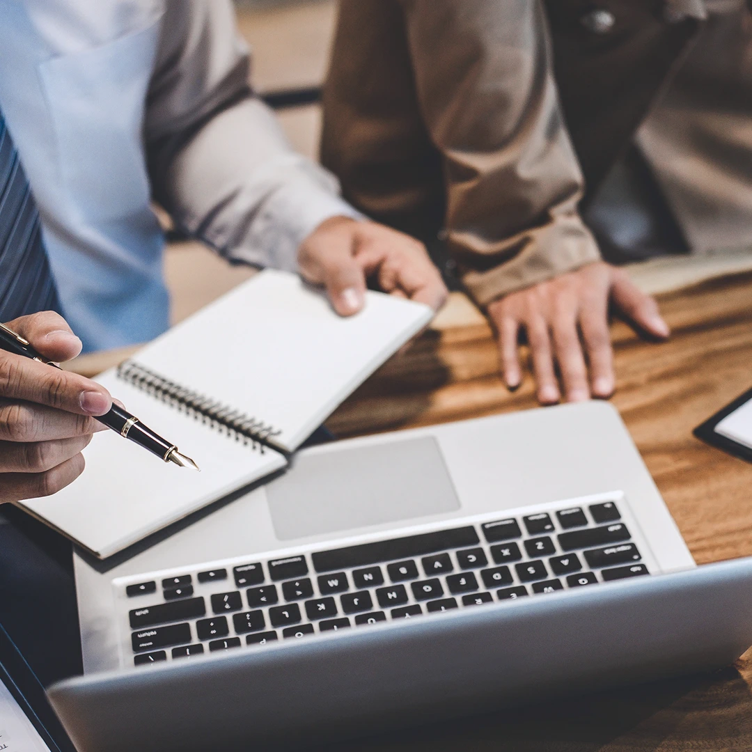 Laptop, notepad, and pen on a wooden desk, with two people working together