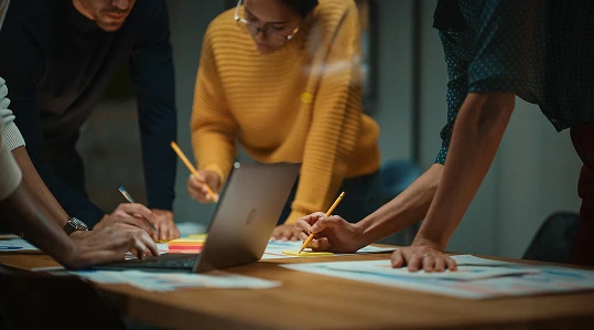 Group of people leaning over a table with documents, sticky notes, and a laptop.