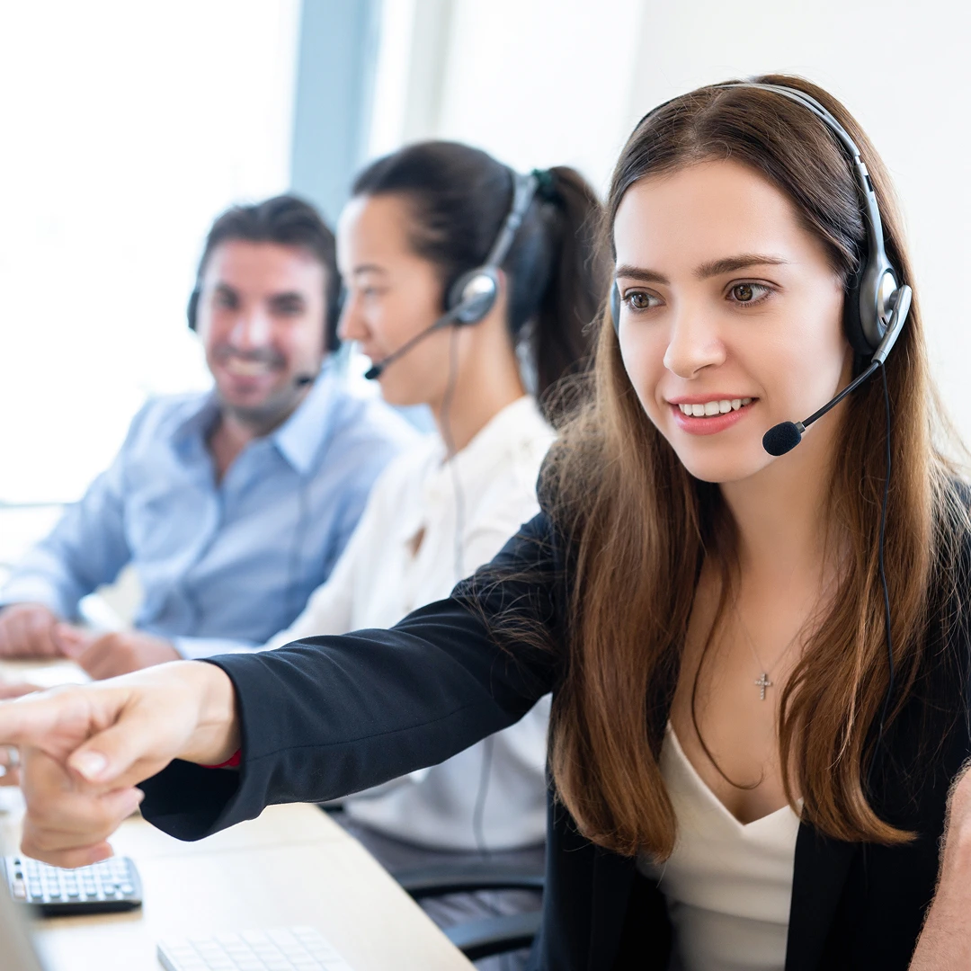 Team of customer service representatives wearing headsets are working at their desks, with a woman in the foreground smiling and pointing at a computer screen