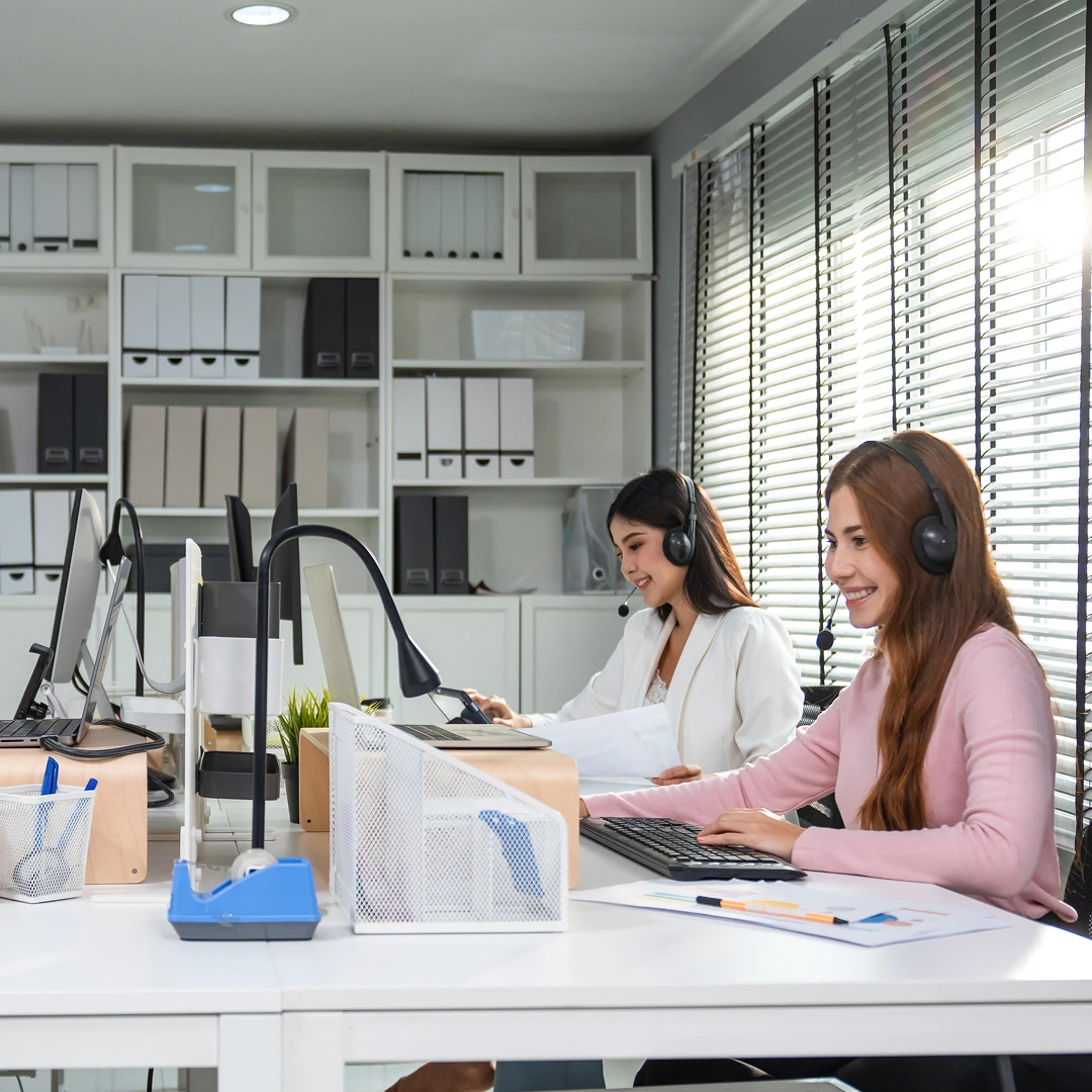 Two female customer support agents wearing headsets, working at computers in an office setting