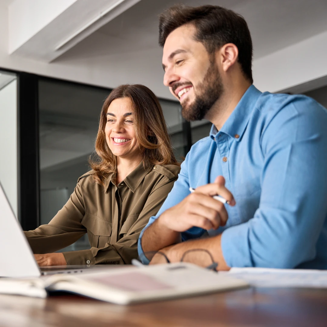 A smiling woman and man sitting at a desk, working together on a laptop in a bright office environment.