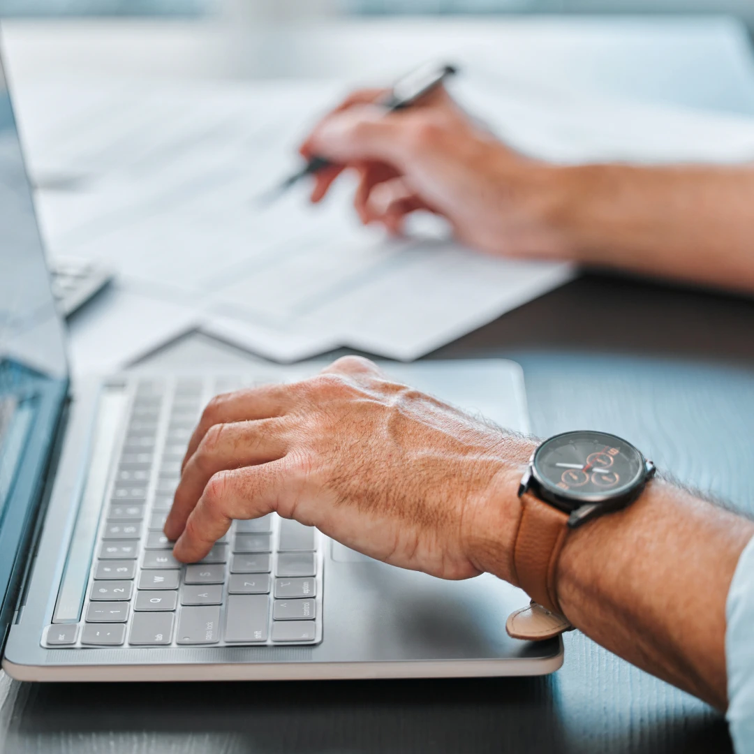 Close-up of hands typing on a laptop keyboard, with documents and a pen behind