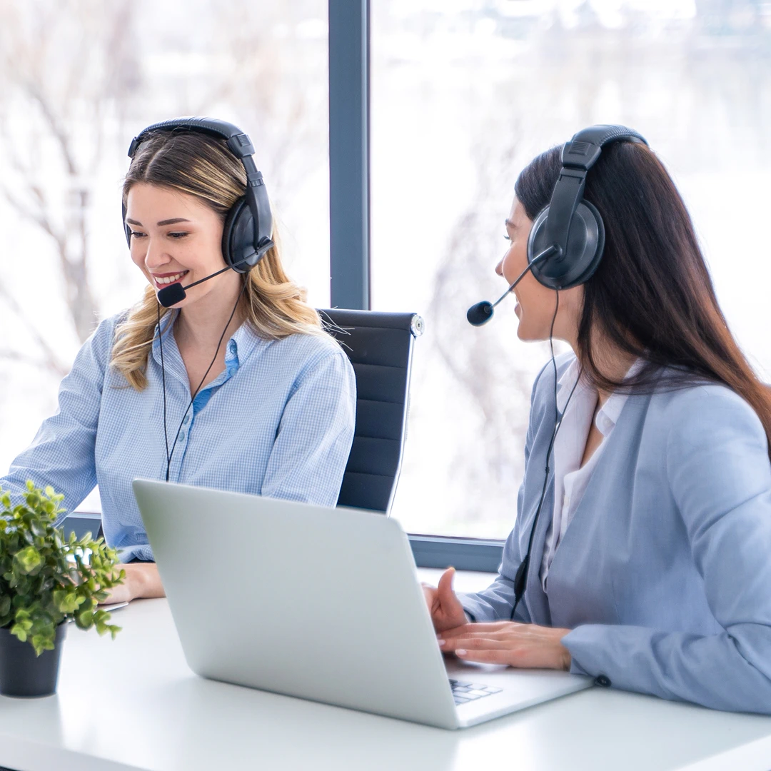Two smiling Polish call center agents wearing headsets and collaborating at a desk with a laptop.