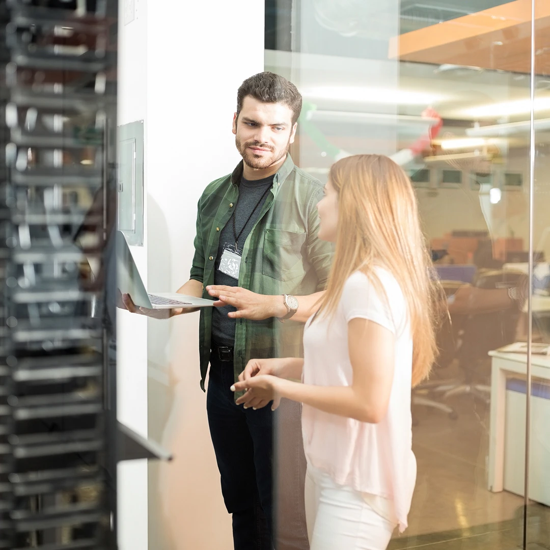 A man with dark hair and a green shirt holding a laptop while talking to a blonde woman wearing a white top in a modern office