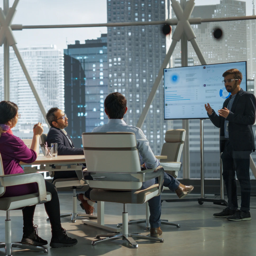 A business meeting in a modern office with a large window, where a man is presenting data on a screen to a group of colleagues seated at a conference table