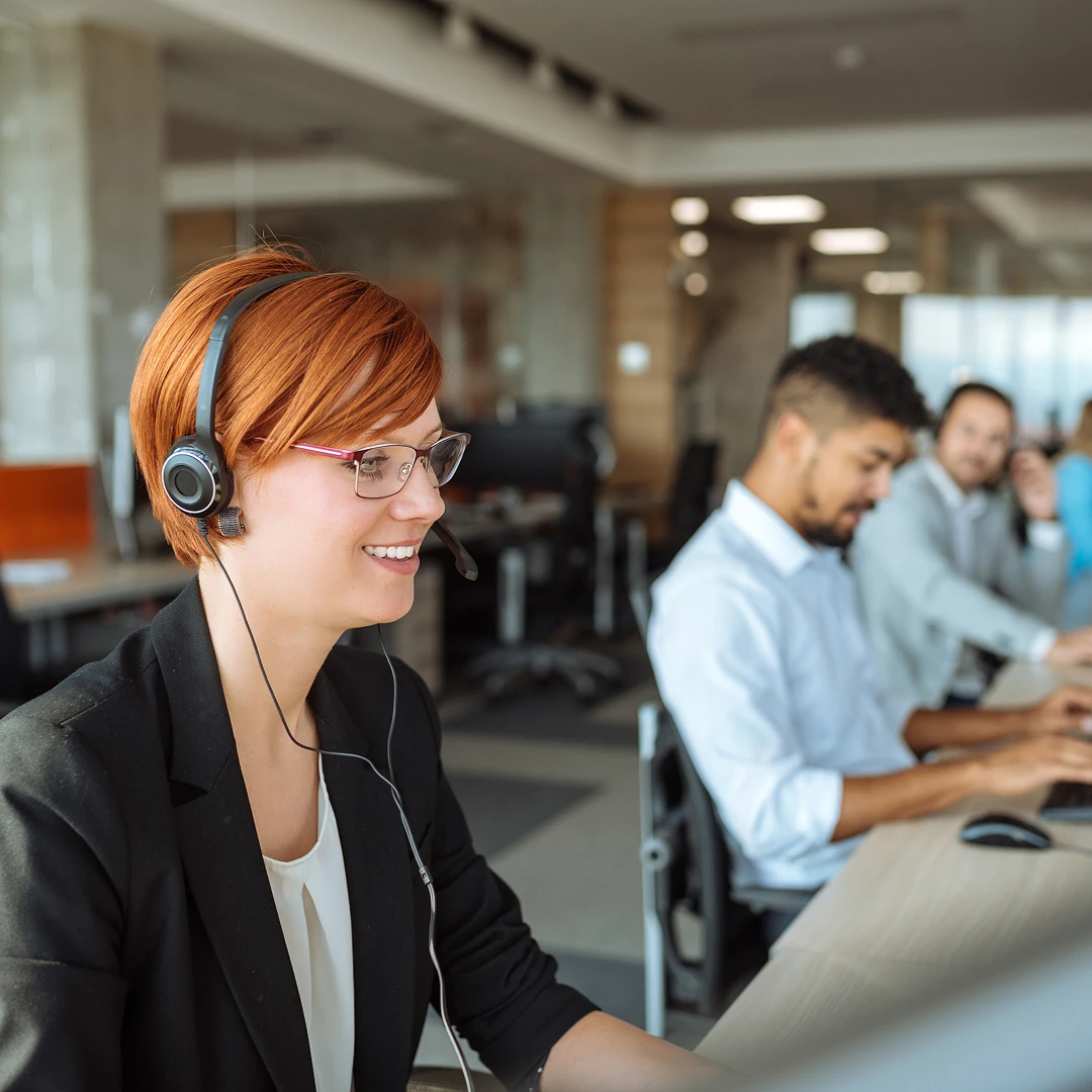 Open office setting with call center agents wearing headsets working at their desks. A woman with red hair and glasses is smiling in the foreground