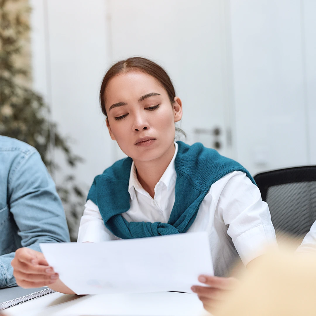 Woman with dark hair and fair skin studying a document at a table, wearing a white button-down shirt with a blue sweater tied around her shoulders