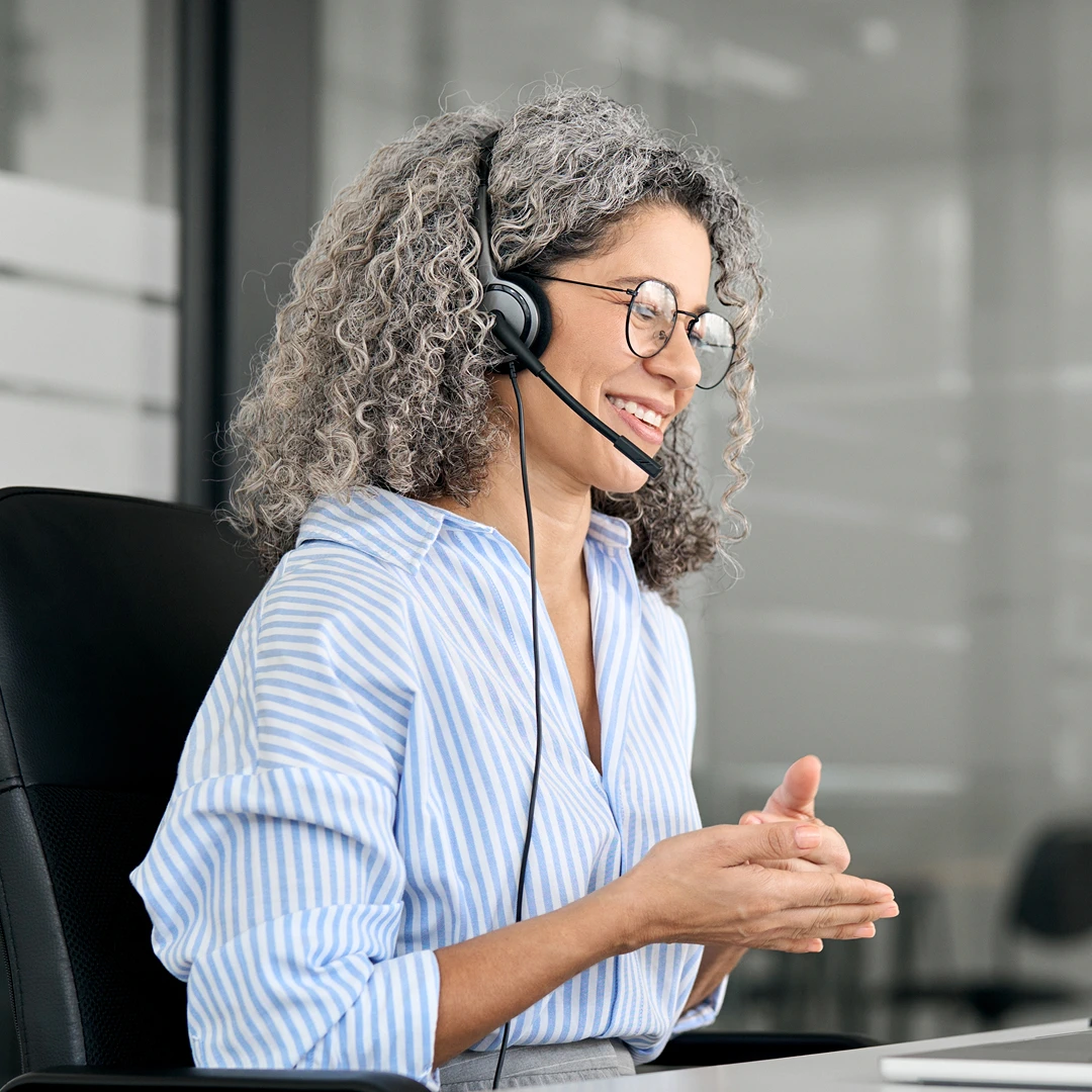 A friendly customer service agent with gray curly hair and glasses speaks into a headset, gesturing with her hands