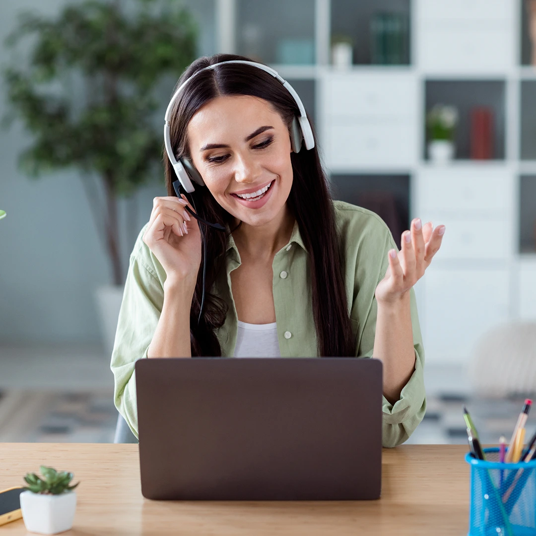 Smiling female financial contact center representative with long brown hair, wearing a headset and light green shirt, gesturing during a video call on her laptop