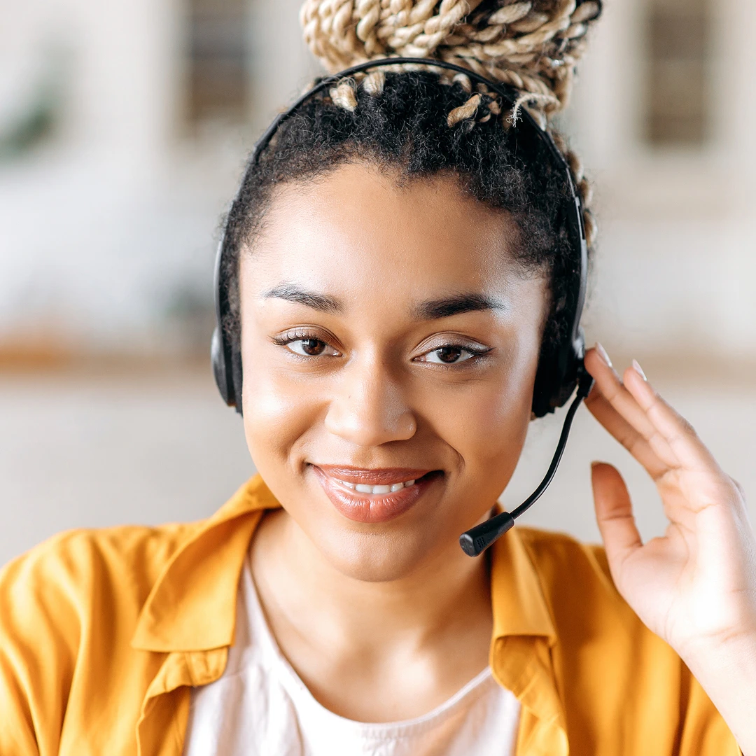 Female customer support agent wearing a headset with a microphone, holding the earpiece with one hand. She has thick braided hair styled in a high bun and is dressed in a bright yellow blouse