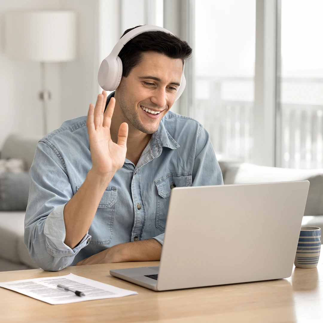 A smiling customer support agent is wearing headphones and waving during a video call on his laptop