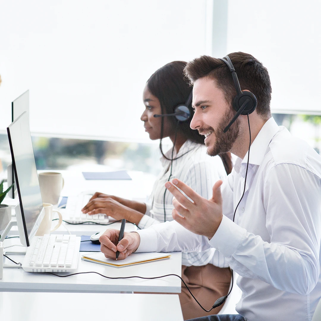 Two customer support agents wearing headsets and white shirts working at their desks, one is writing in a notebook while speaking