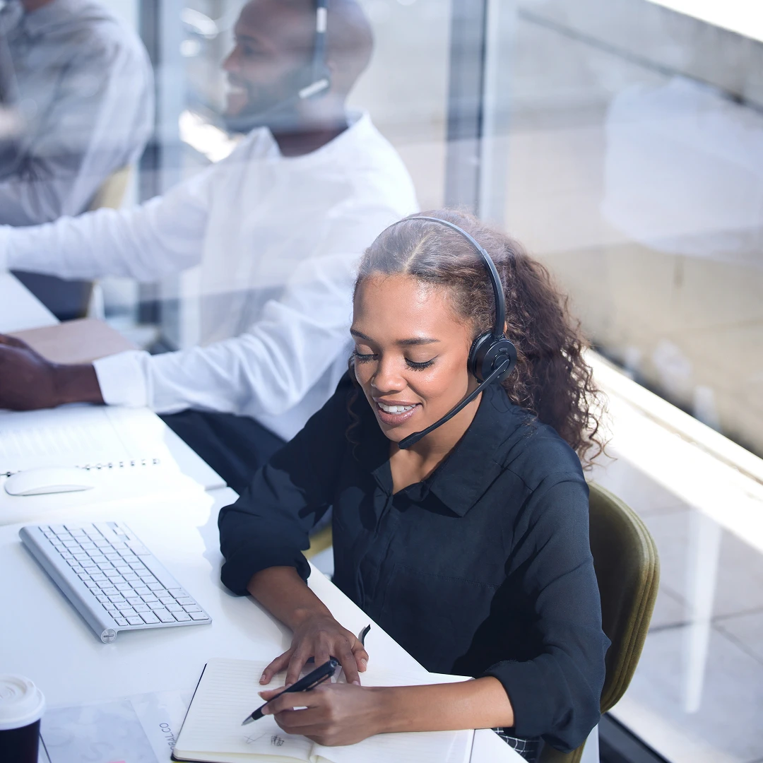 Smiling woman with curly hair working at a desk, wearing a black blouse and headset, and writing in a notebook