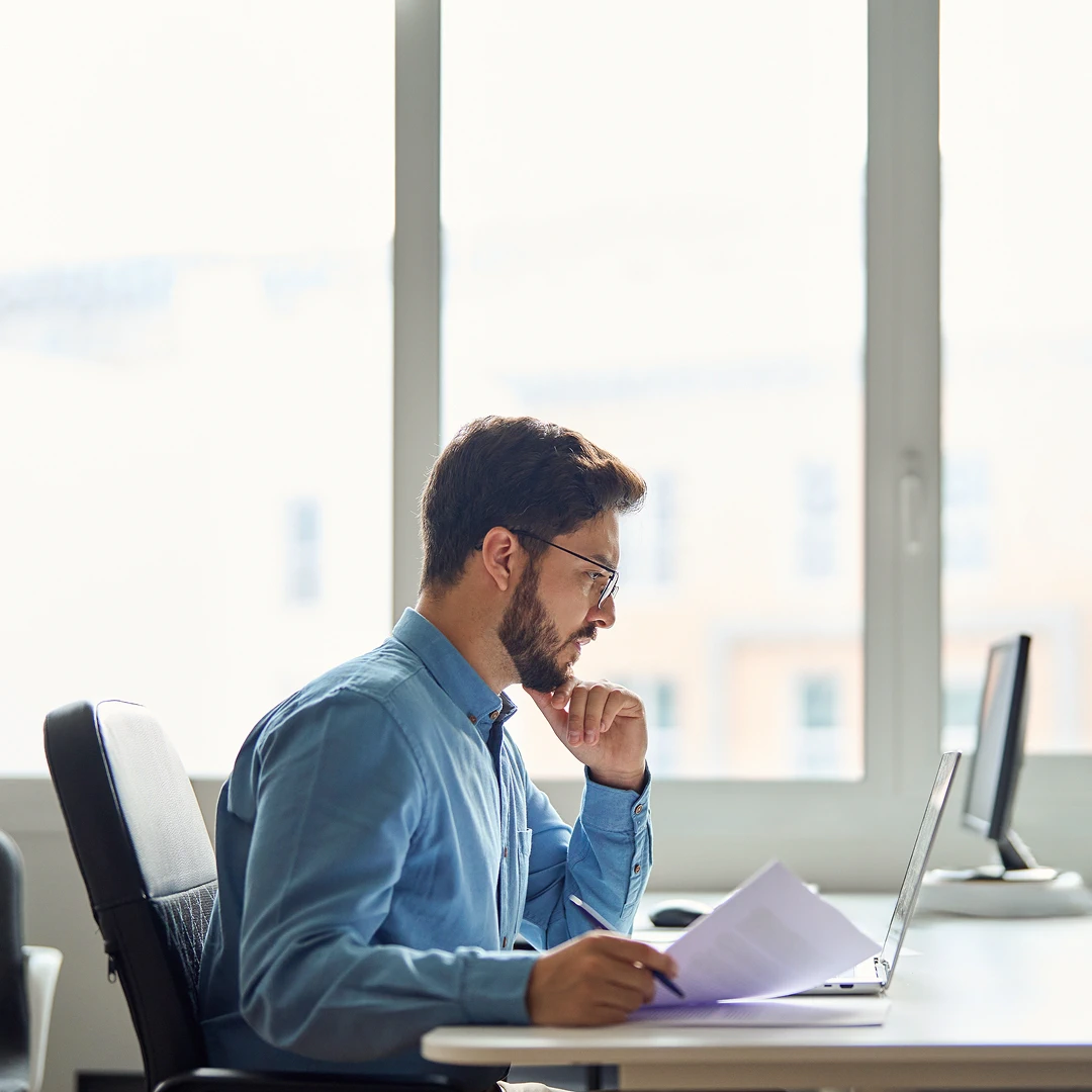 Man with beard and glasses working at a desk with a laptop and documents