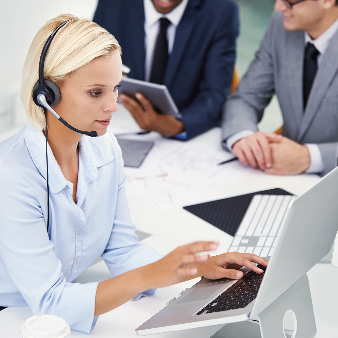 Blonde female customer service representative wearing a white blouse and headset, assisting customers and working on a laptop
