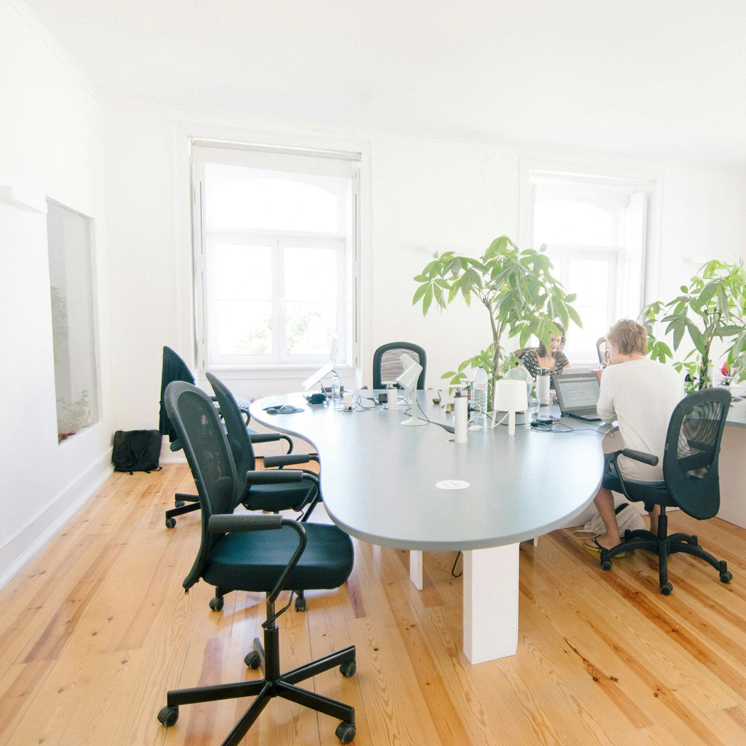 Bright, modern office with a large conference table, several plants, and two people working on laptops