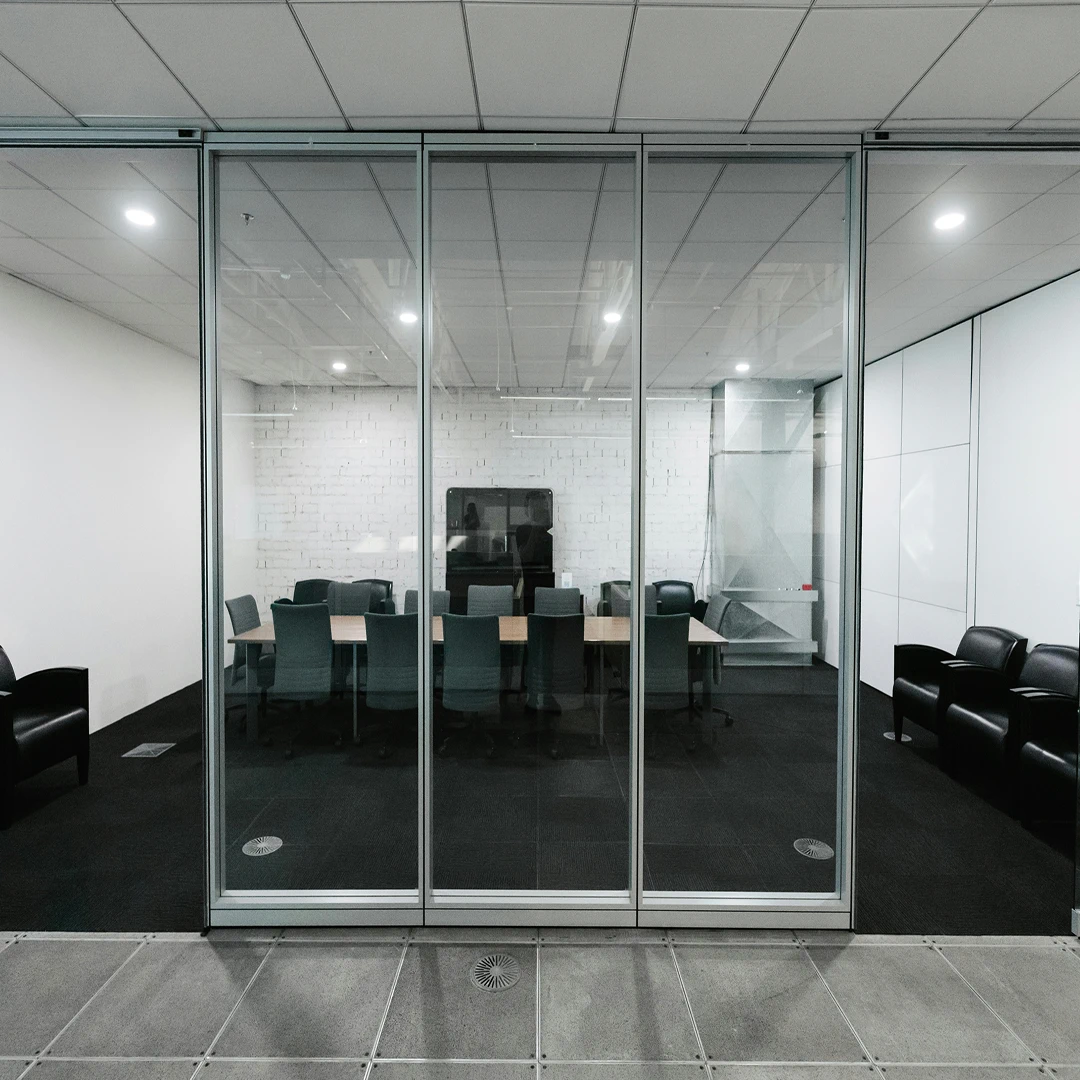 Bright conference room with a rectangular table, gray office chairs, and a floor-to-ceiling glass wall on one side