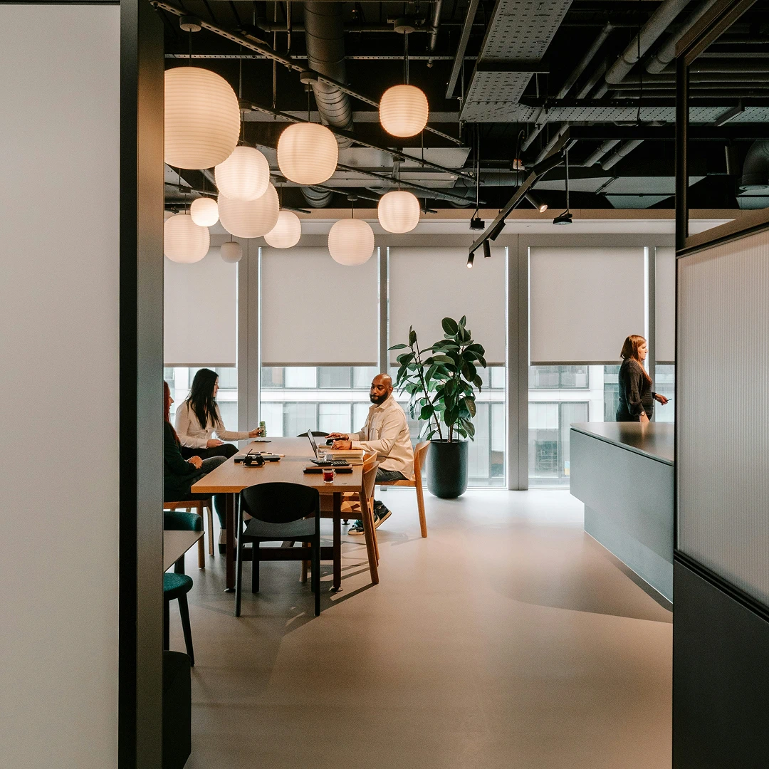 Modern office interior featuring a group working at a table, suspended paper lanterns, large windows, and a green plant