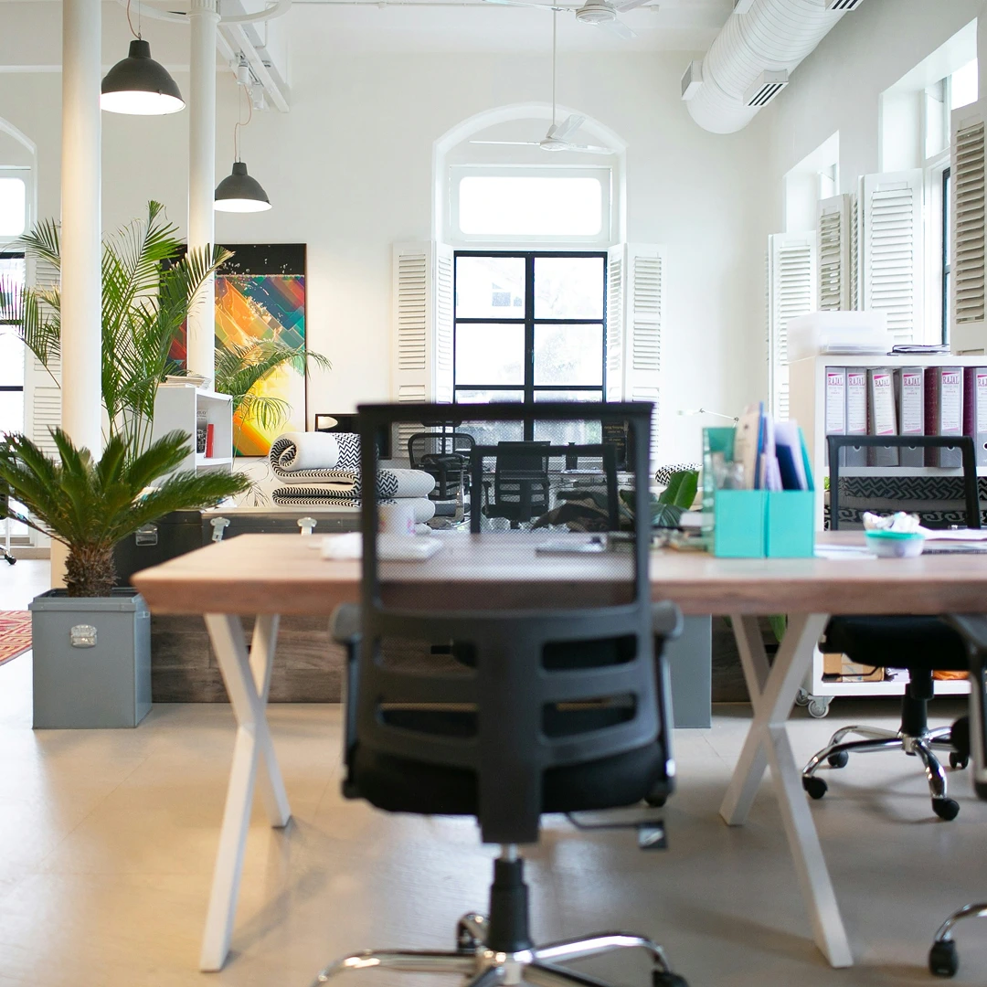 A modern, well-lit office featuring wooden desks, black chairs, and large windows