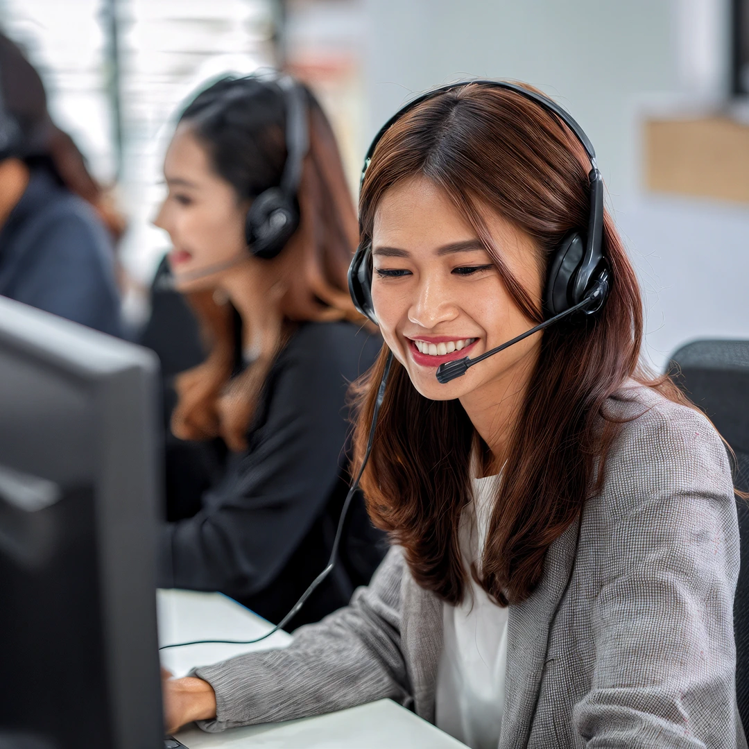 A smiling woman with long dark hair, wearing a gray blazer, is working in a busy Filipino call center