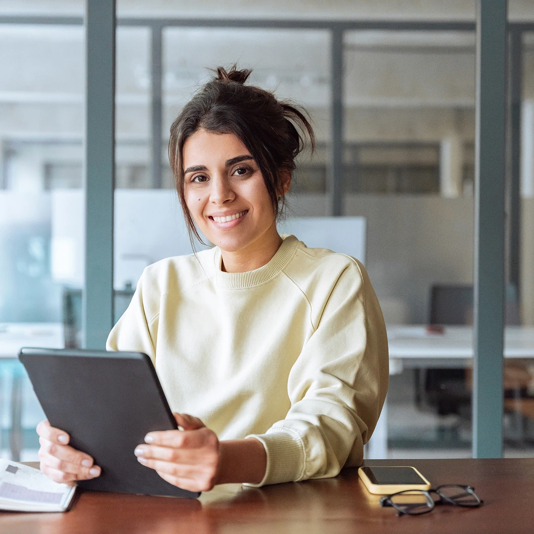 A smiling woman wearing a loose yellow sweater, sitting at a desk and holding a tablet in her hand