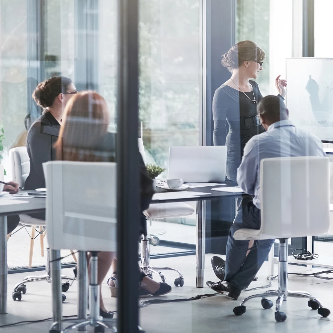 Team meeting in a modern glass-walled office, with a woman presenting to colleagues