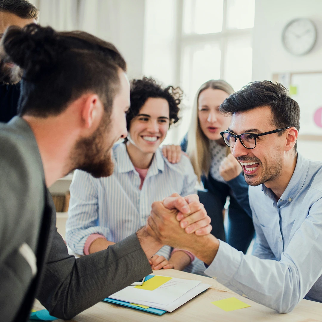 Business colleagues having fun with an arm wrestling match in the office, with supportive team members cheering them on