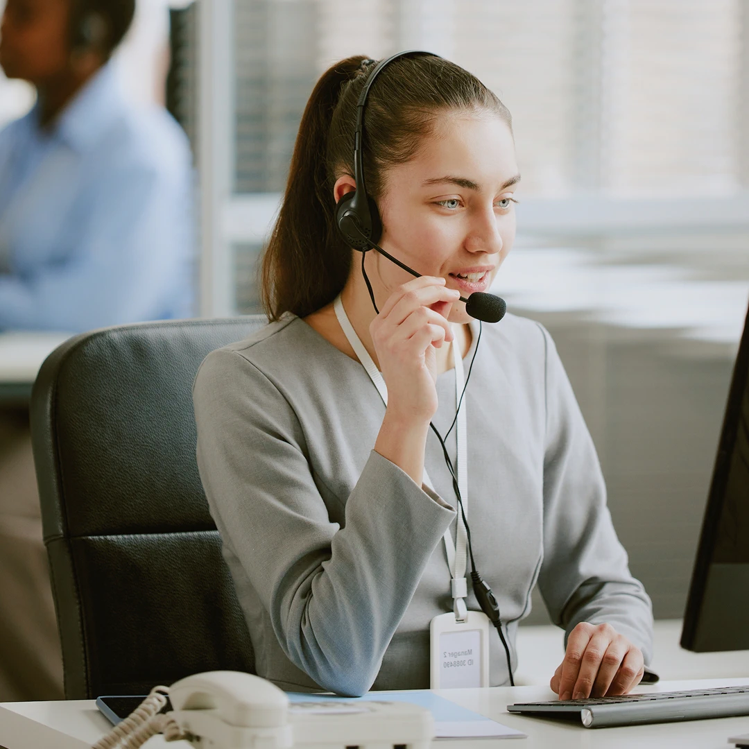 Friendly female customer support agent wearing a grey suit with a headset in a bright office working at a computer