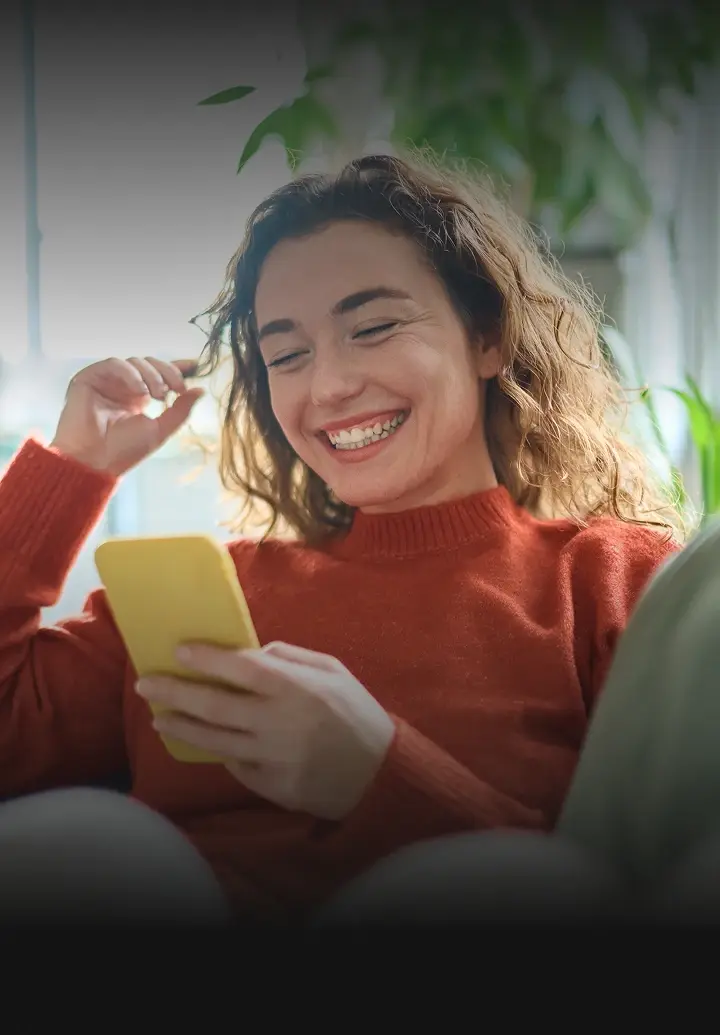 A smiling woman with curly hair, wearing a red sweater, looks at a yellow smartphone she is holding.