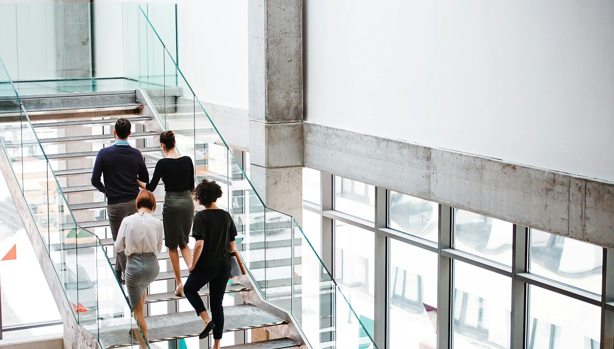 Modern office staircase with glass railings, large windows, and employees walking upwards