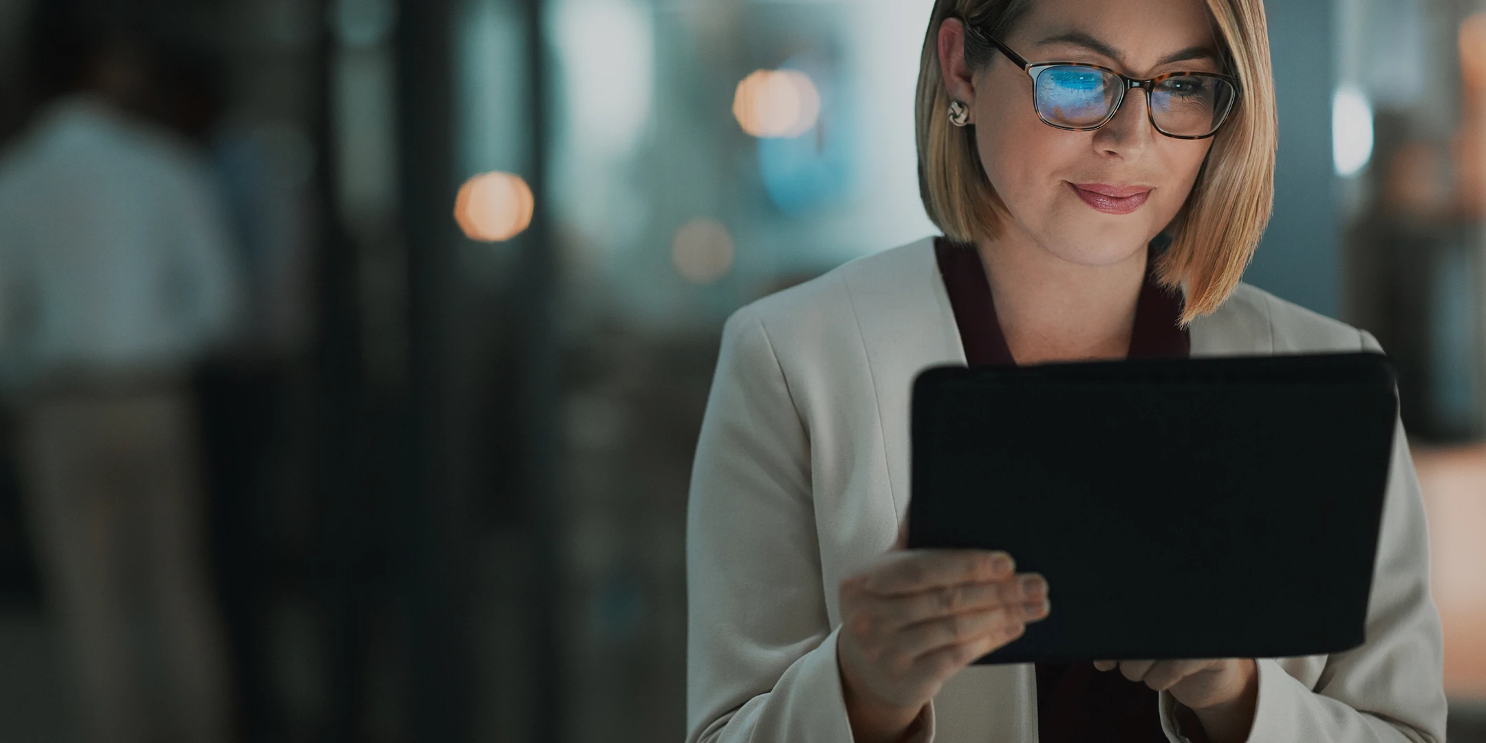 A woman with short blonde hair and tortoiseshell glasses, wearing a cream-colored blazer, working on a tablet