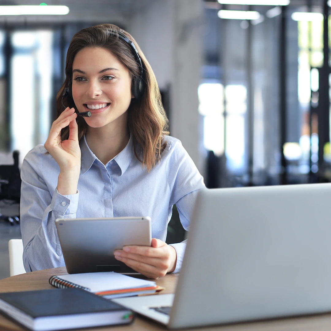 A cheerful female customer support representative wearing a blue blouse and headset, working at her workstation in a modern office in Ukraine.