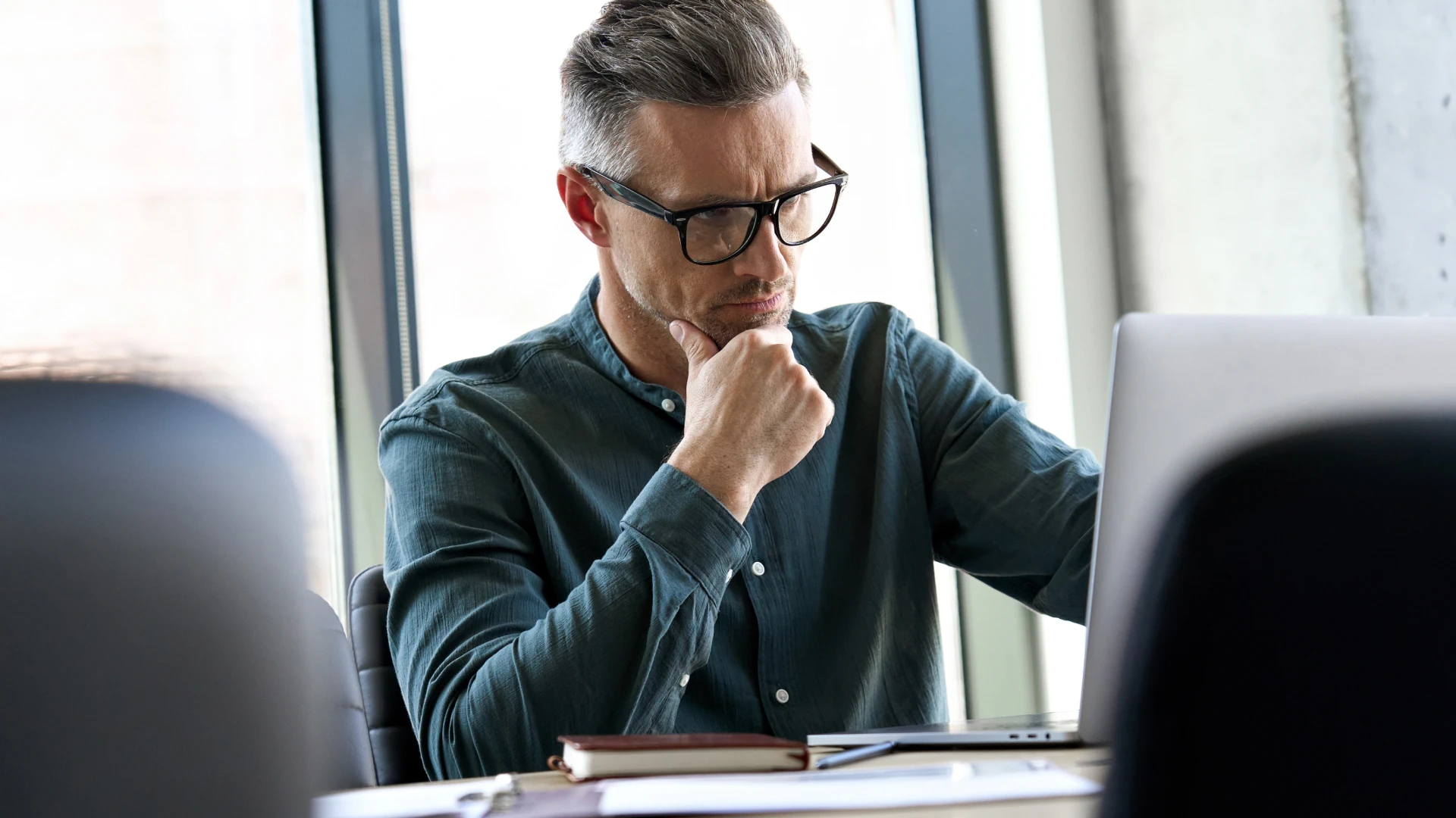 A man in a gray shirt and glasses is sitting in front of a laptop, thoughtfully resting his chin on his hand