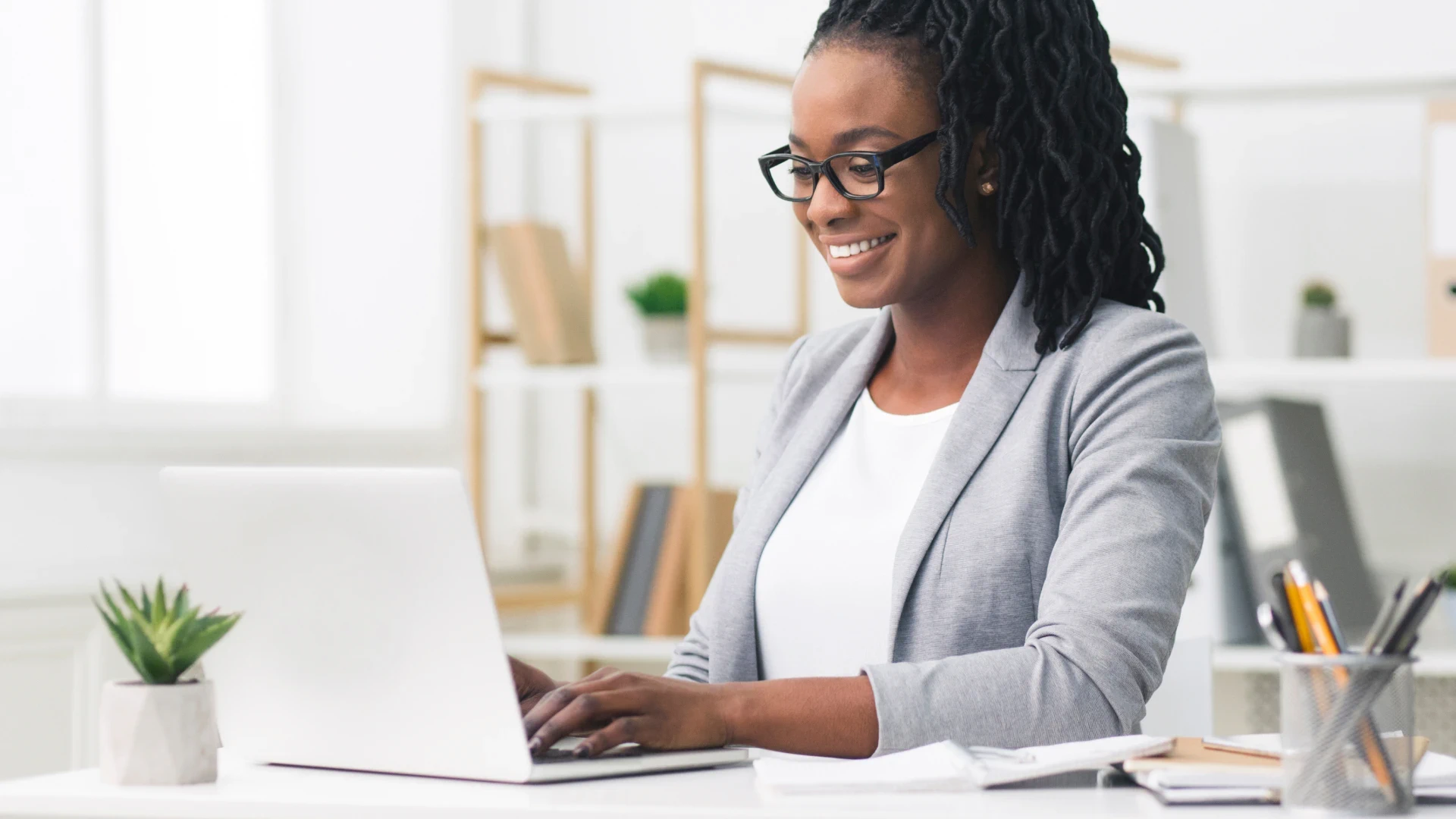A professional woman wearing glasses and a gray blazer is seated at a desk, smiling as she works on a laptop in a brightly lit office
