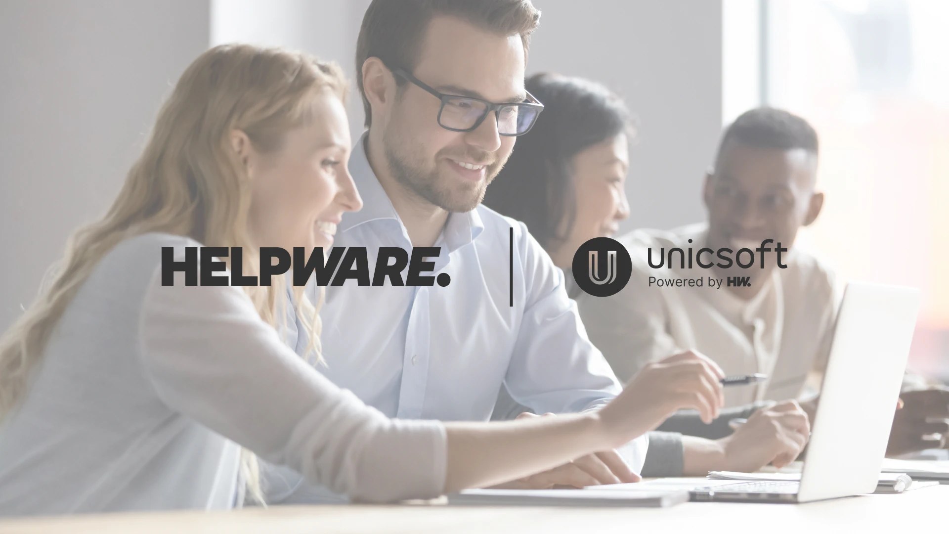 A group of four people sitting together at a table, smiling and collaborating while working on laptops and documents in a bright, modern office