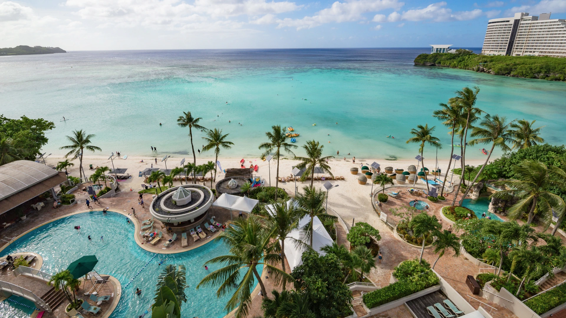 Aerial view of a beachfront resort with palm trees, a pool area, and white sand leading to turquoise ocean water