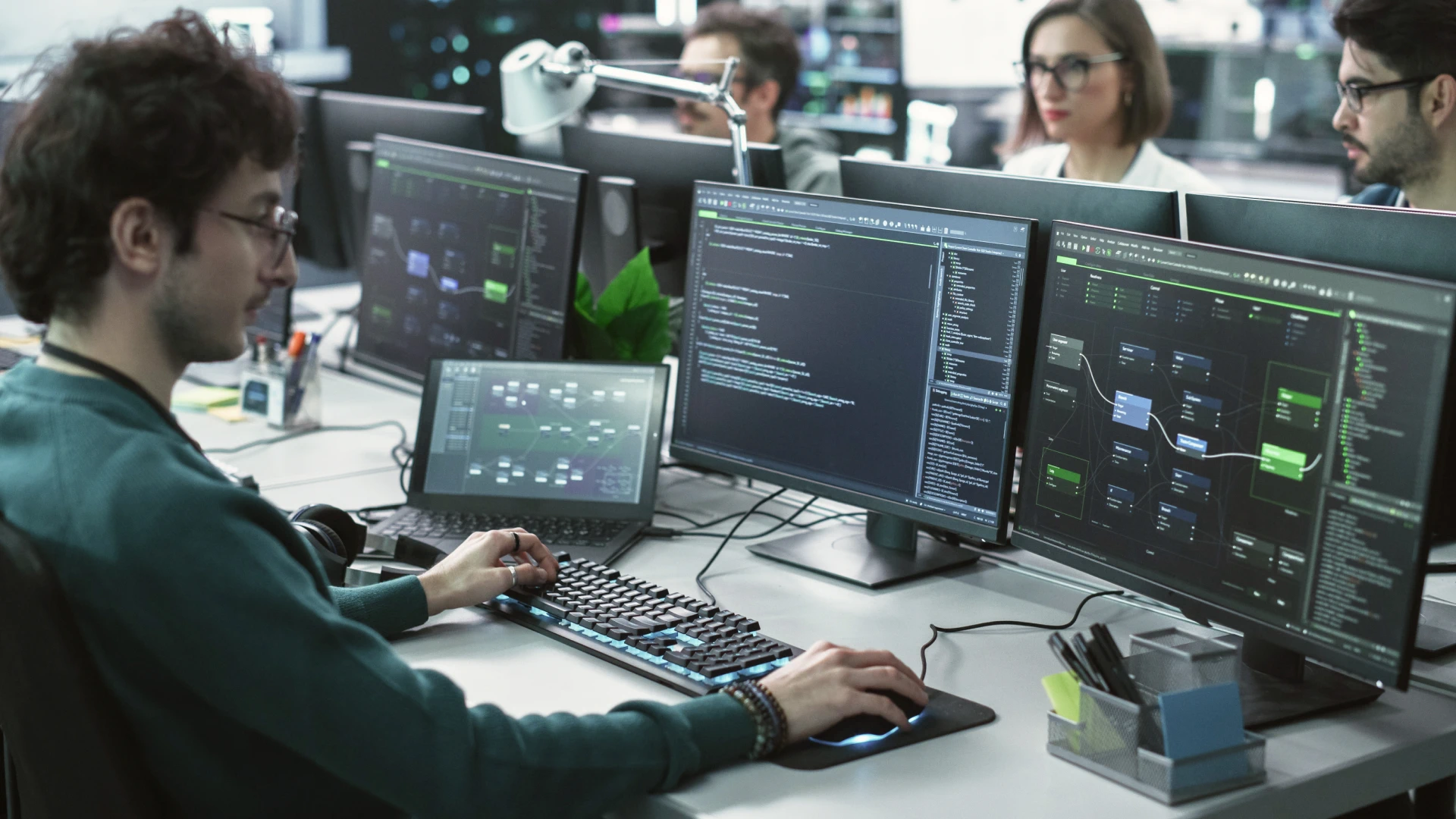 A man in glasses, wearing a green sweater, is typing on a keyboard in a modern office setting, surrounded by multiple computer screens