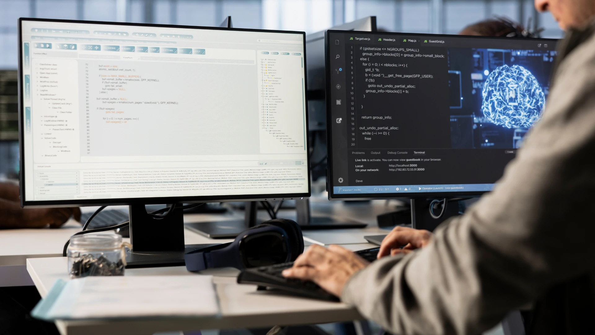 A man is sitting at a desk, working on a computer with two monitors, and documents and headphones are lying on the desk