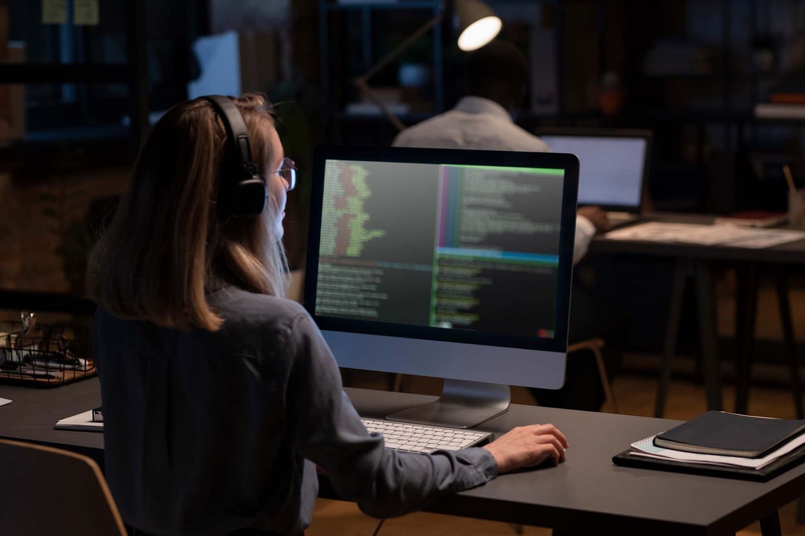 A woman wearing a grey shirt and glasses is working at a desk on a computer in a large open-space office.