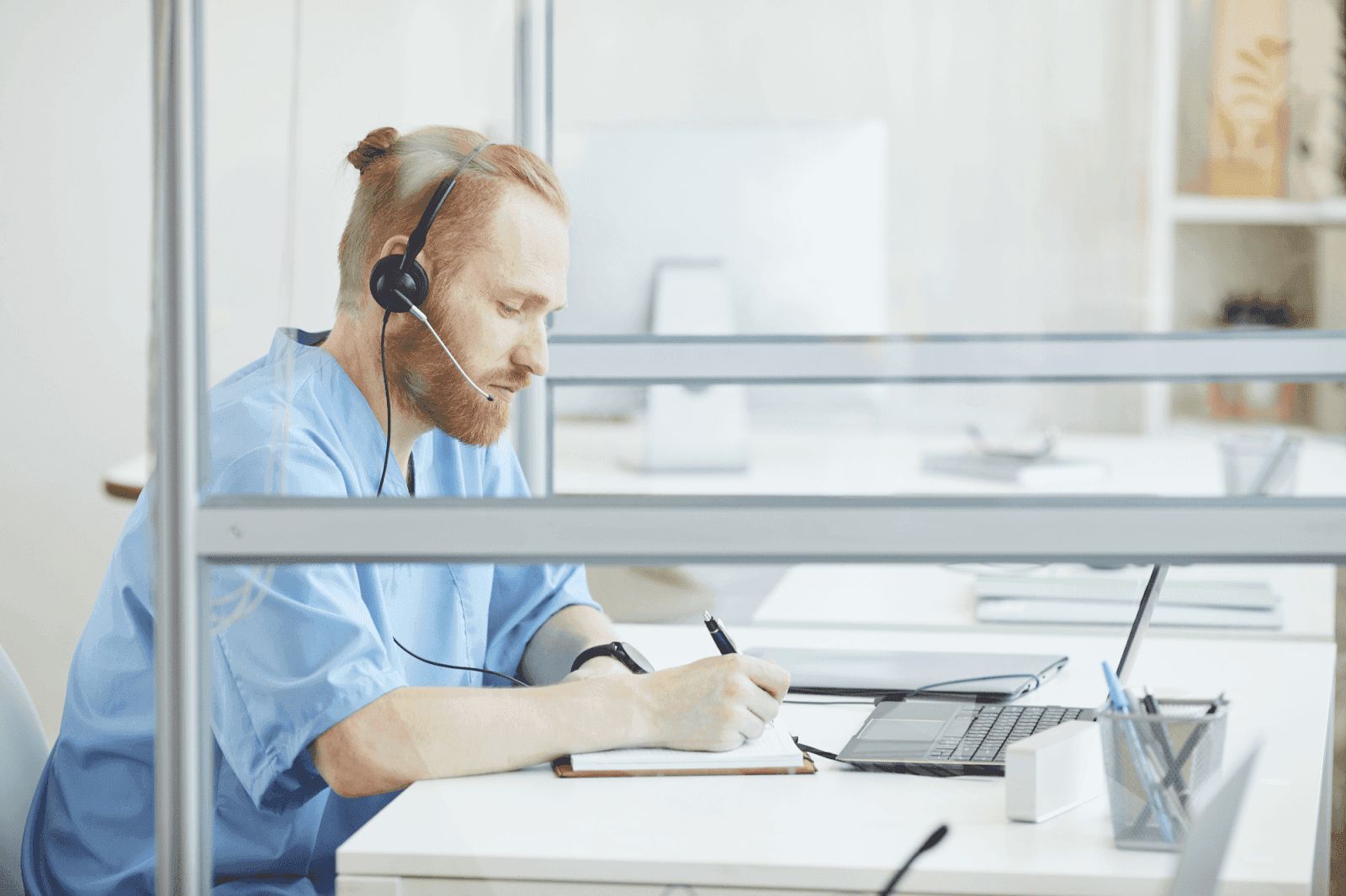 A customer service agent wearing a headset is working at a laptop and taking notes in a notebook.